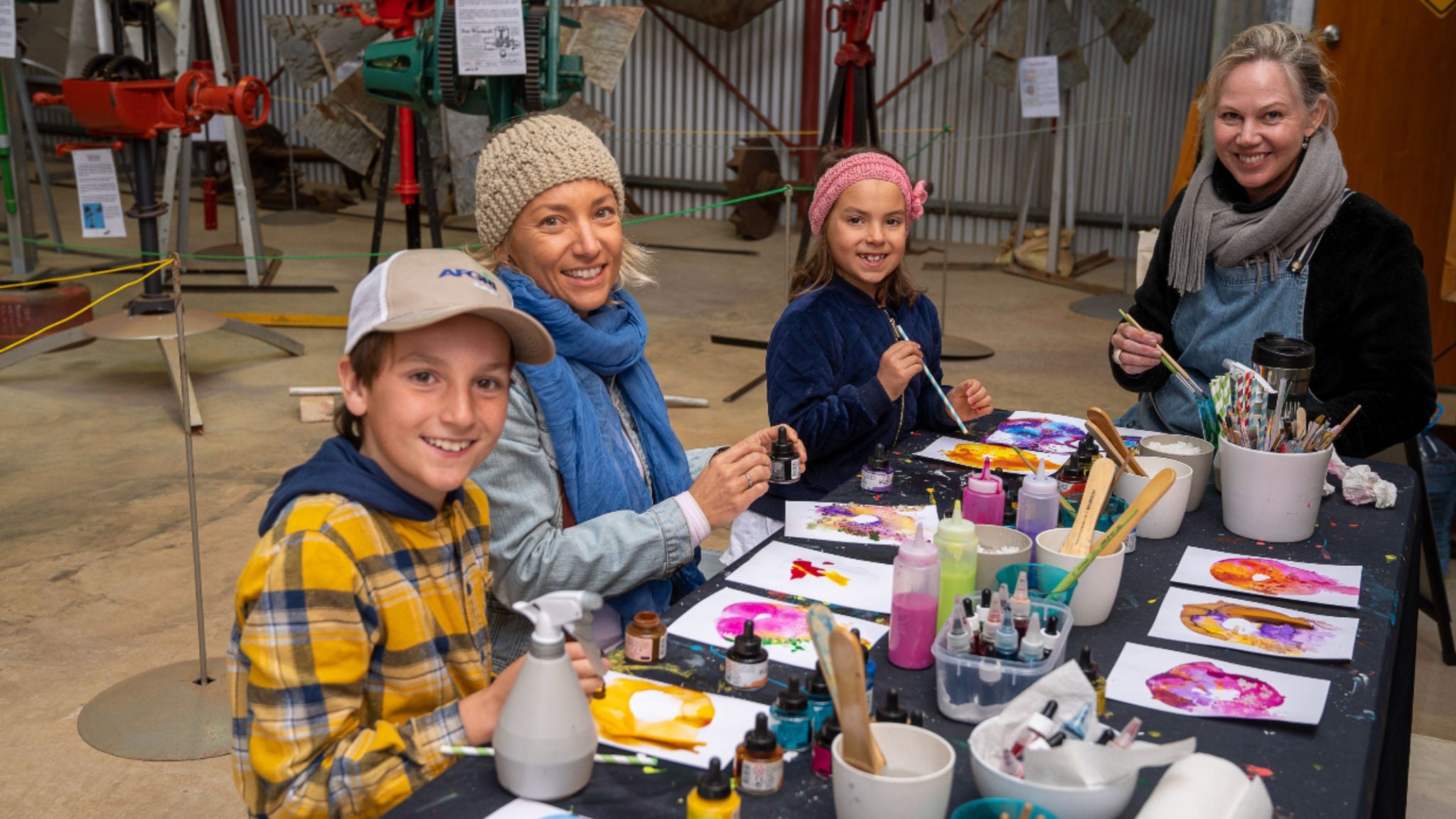 A group of children and adults are seated around a table, engaging in a creative painting activity. The setting appears to be an indoor workshop or studio, with various art supplies such as paint bottles, brushes, and paper visible on the table. The participants are focused on their colorful artwork, which features vibrant hues and abstract designs. The background includes industrial equipment, suggesting a community or educational venue.