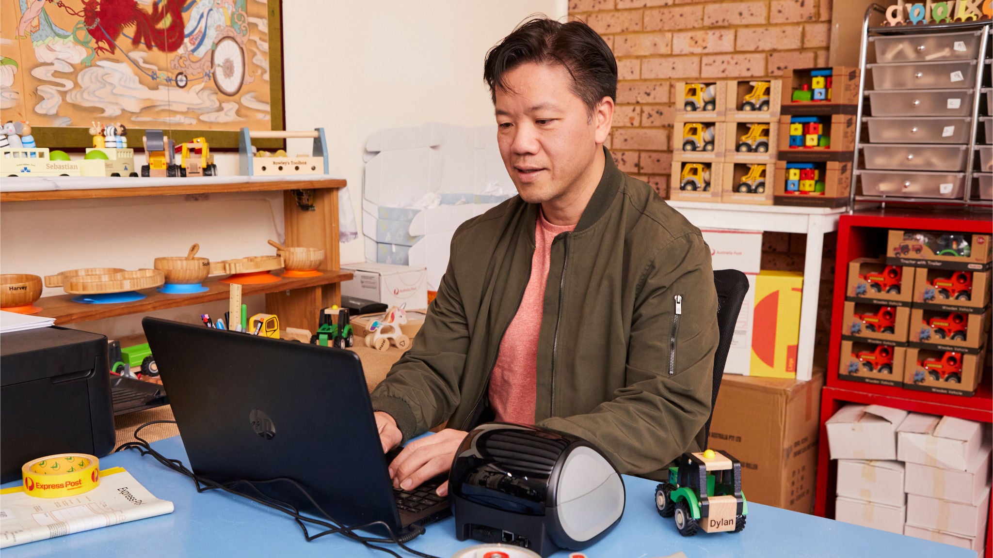 An adult male sits at a blue desk using a laptop, surrounded by various office and toy items. The setting appears to be a creative workspace or classroom, with shelves of organized supplies and toy vehicles visible. The atmosphere is casual and vibrant, with a mix of educational and playful visuals. A label printer and a green toy truck are prominent on the desk, along with a roll of yellow tape marked 'DYMO'.
