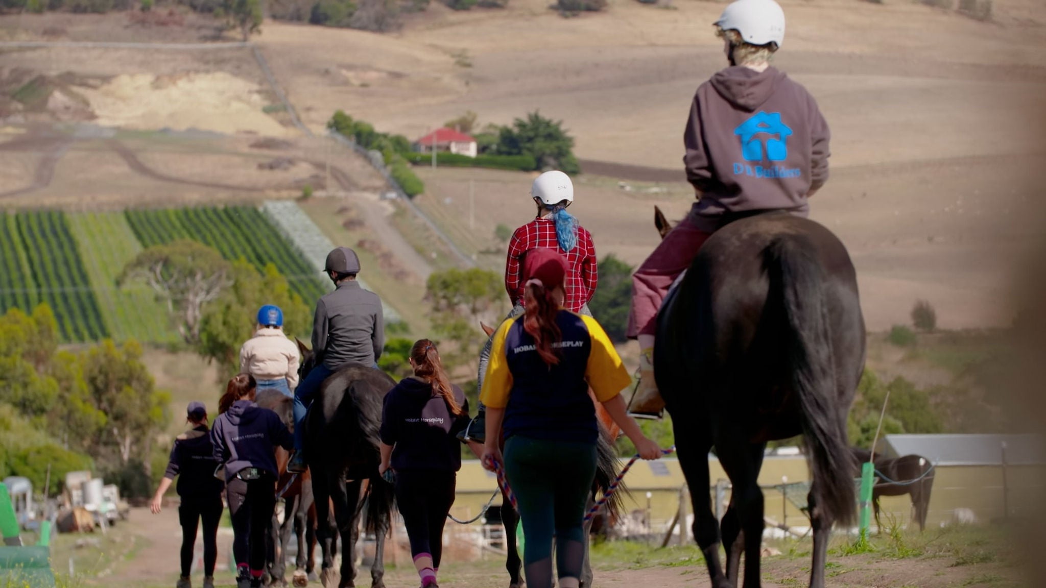 A group of people, including adults and children, ride horses along a rural trail accompanied by instructors. The riders wear helmets and casual outdoor clothing, and the scene overlooks farmland and rolling hills. The mood is relaxed and recreational, with earthy tones dominating the landscape. No visible text or numbers are present in the image.