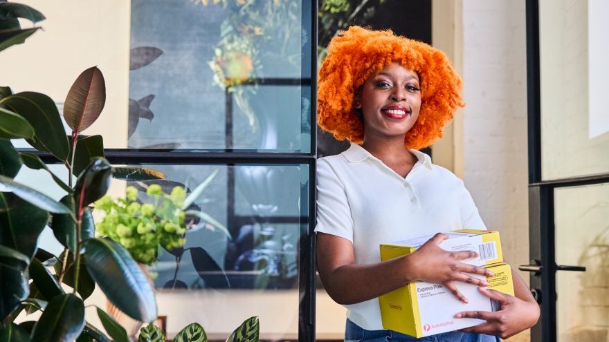 A young woman with bright orange curly hair smiles while holding several yellow and white boxes. She stands in a modern indoor space with large glass panels and a black-framed door. Green plants and soft natural light create a fresh, contemporary atmosphere. The scene suggests parcel delivery, online shopping, or small business shipping.
