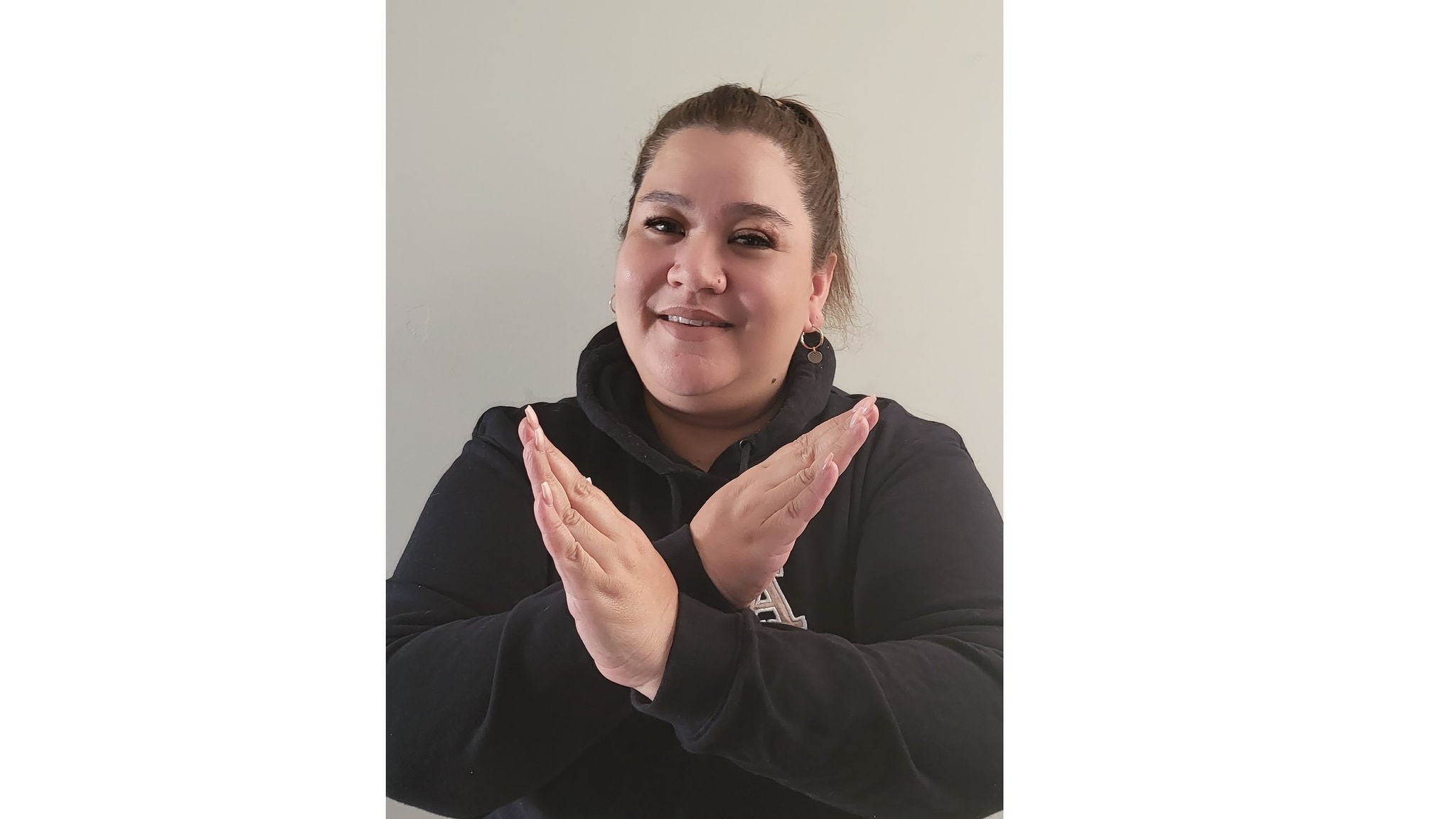 A person is pictured indoors with their arms crossed in front of their chest, forming an 'X' gesture. The individual is wearing a dark hoodie and is positioned against a plain, light-colored wall. The gesture suggests a signal of refusal, stop, or disagreement. No visible text, logos, or numbers are present in the image.