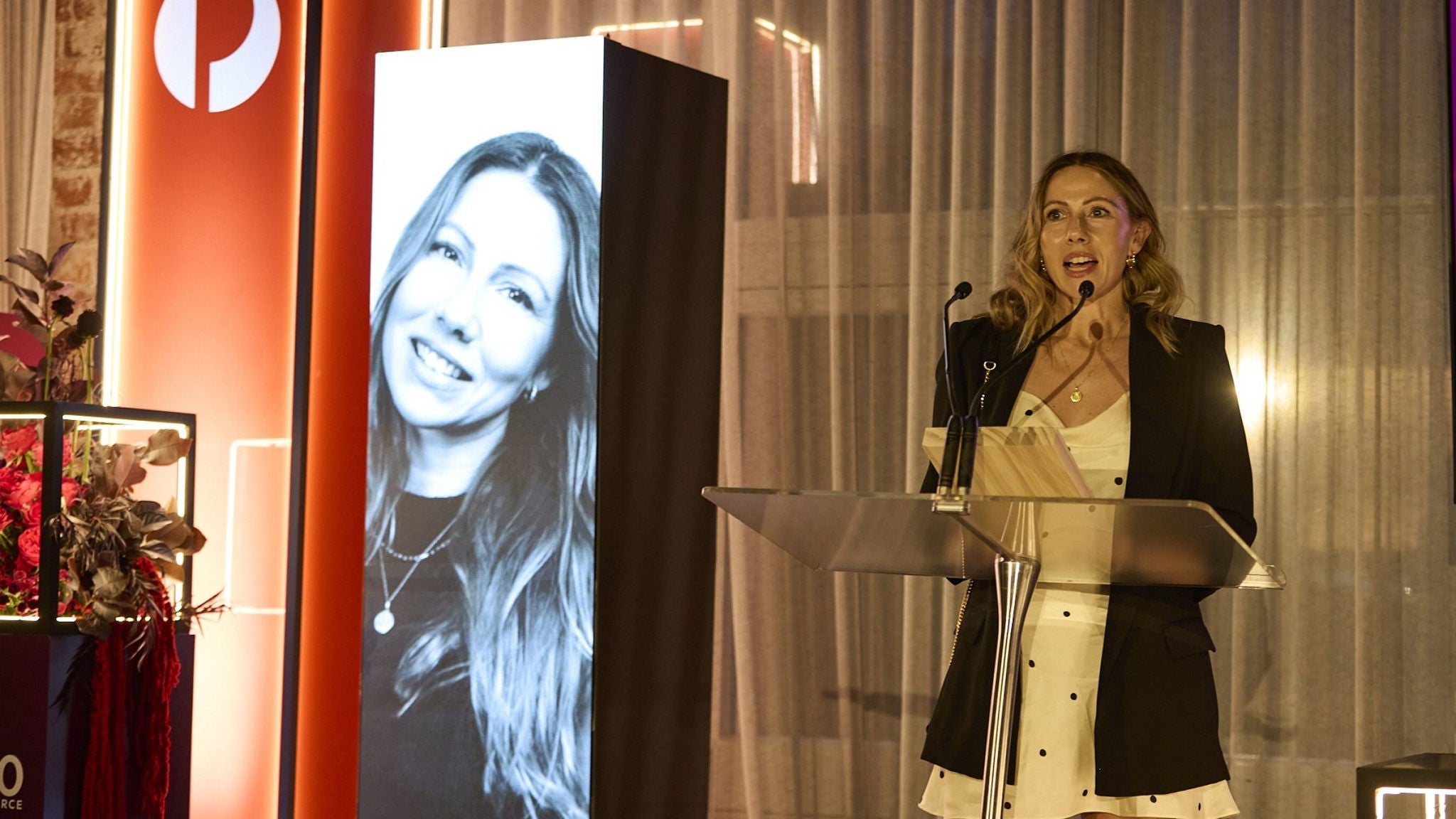 A woman stands at a clear podium giving a speech at an indoor event. Behind her, tall illuminated panels and a large vertical screen with a portrait image create a dramatic backdrop. Warm lighting and sheer curtains contribute to an elegant atmosphere. Floral arrangements and modern display structures suggest a formal ceremony or corporate celebration.