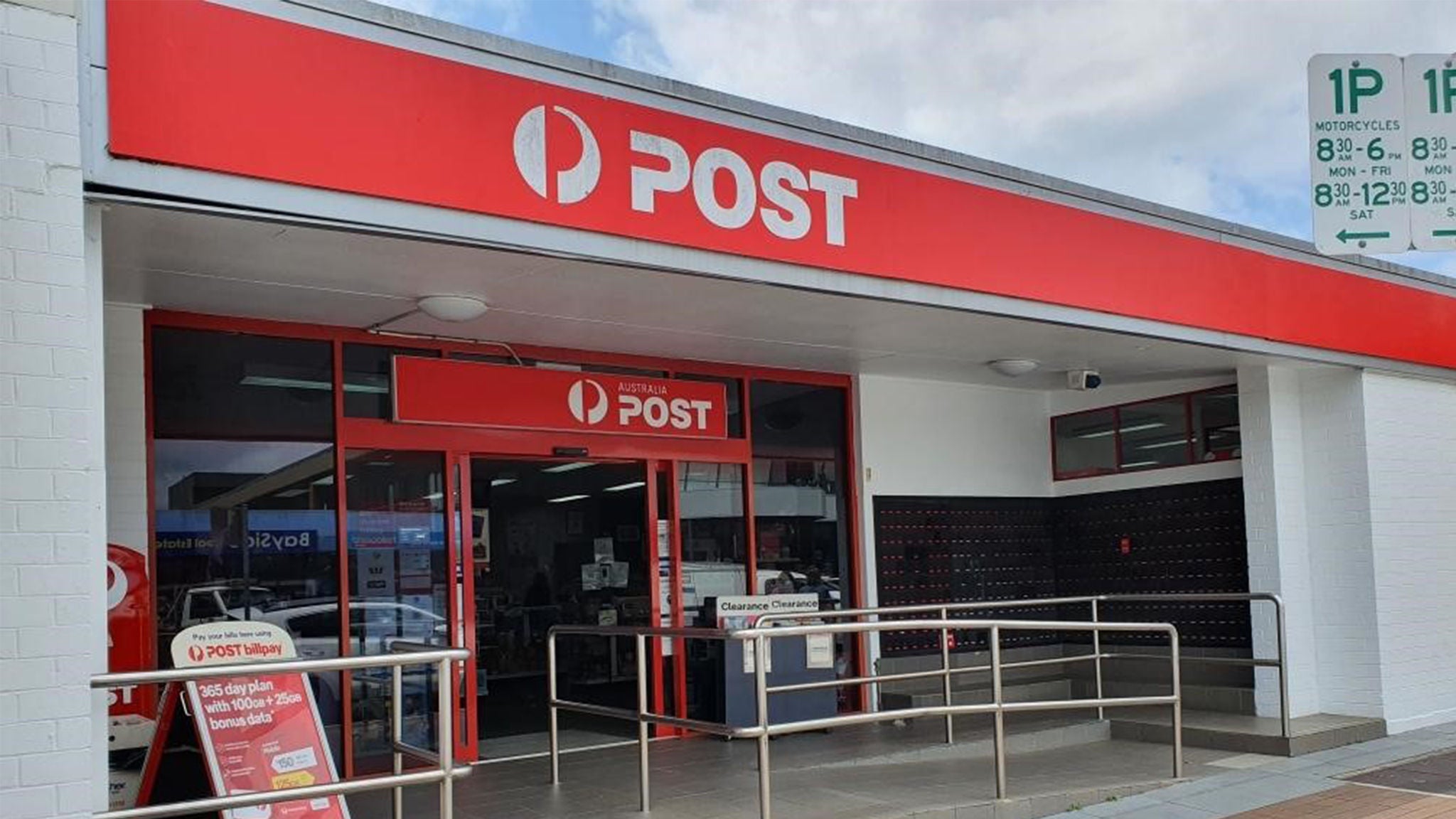 The image shows the exterior of an Australia Post office, featuring prominent red and white signage with the word 'POST' clearly visible. The entrance includes glass doors, accessibility ramps, and a display board with service information. A parking sign with the numbers '1P', '8:00-6:00', and '8:12-12:00' is visible above the entrance. The setting is urban, with a clean and functional appearance.