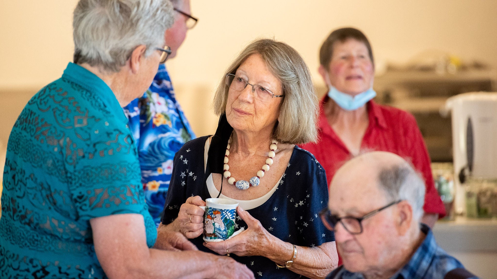 A group of older adults are seen conversing in a warmly lit indoor setting. One woman holds a decorative mug and wears a large beaded necklace, while others are engaged in friendly conversation. The atmosphere appears casual and welcoming, with participants dressed in colorful attire. The background suggests a community or recreational space. No visible text or numbers are present in the image.