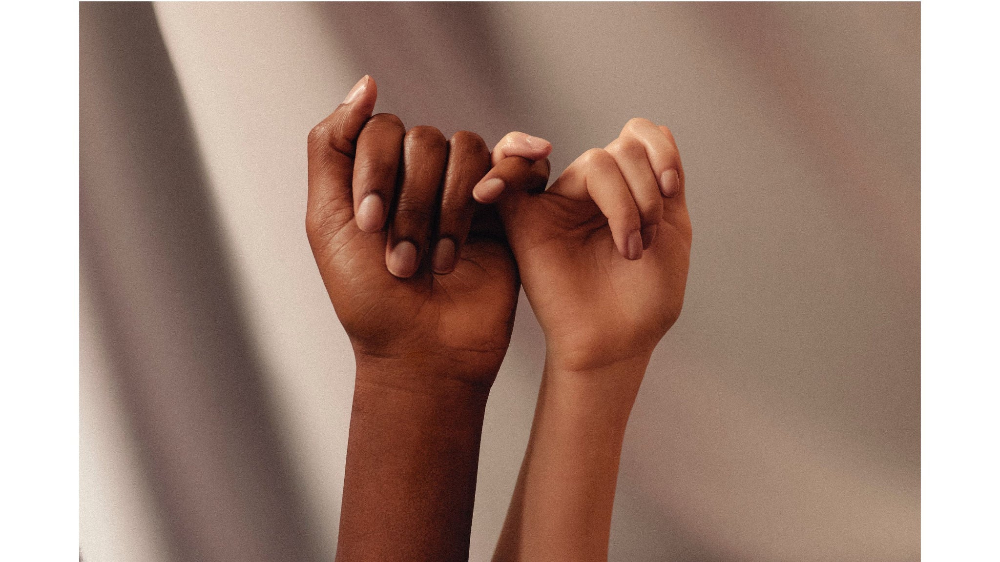 The image features two hands of different skin tones interlocking their pinky fingers in a pinky promise gesture. The background is softly blurred with neutral, warm tones, creating a calm and harmonious atmosphere. The focus is on the hands, highlighting themes of unity, trust, and diversity. No visible text or numbers are present in the image.