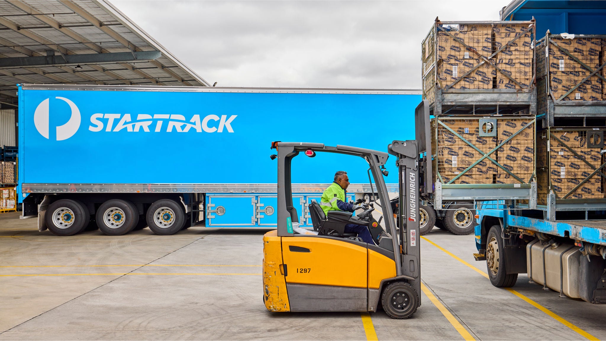 A worker operates a yellow forklift, transporting a pallet of goods beside a large blue StartTrack truck trailer.