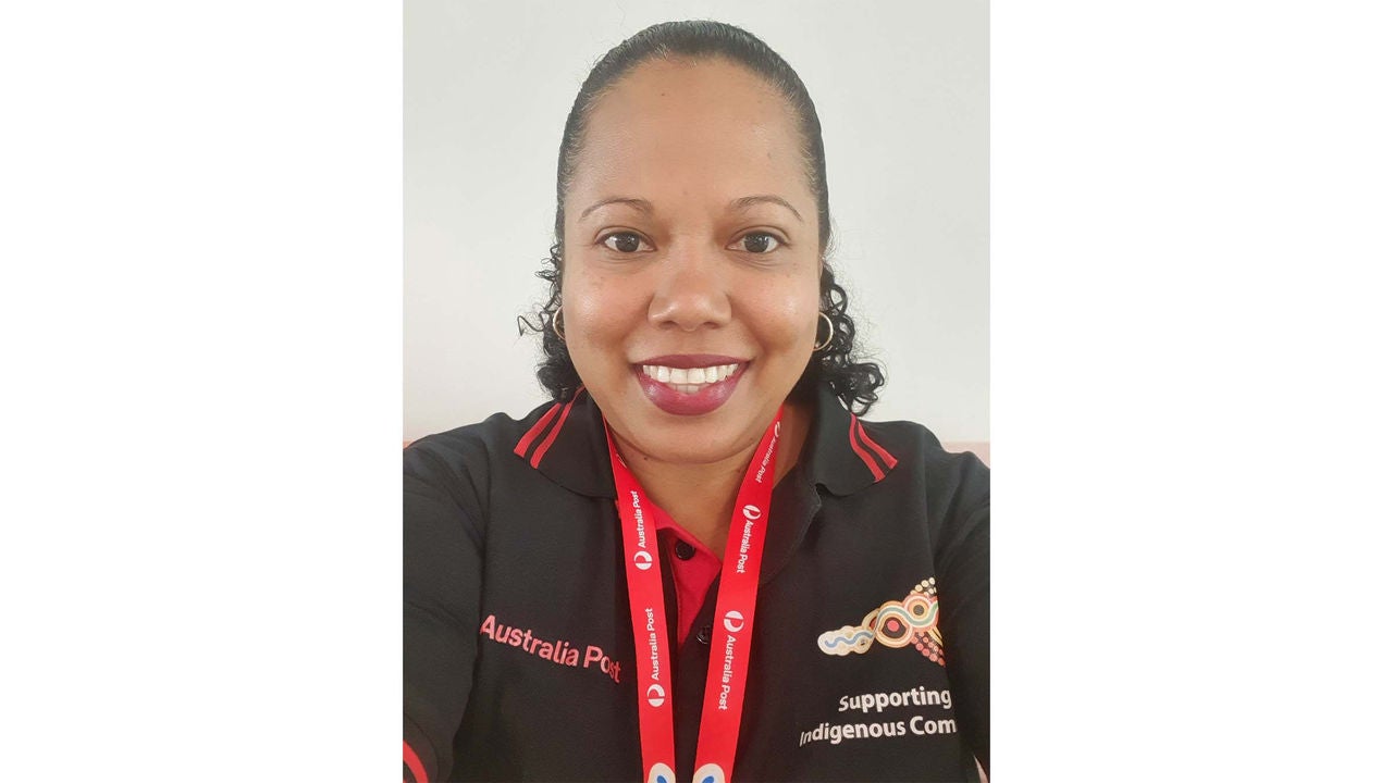 A person is pictured wearing a black Australia Post polo shirt with red accents and a red lanyard. The shirt features text supporting Indigenous communities and a distinctive design on the chest. The background is plain and neutral, focusing attention on the uniform and visible text elements.