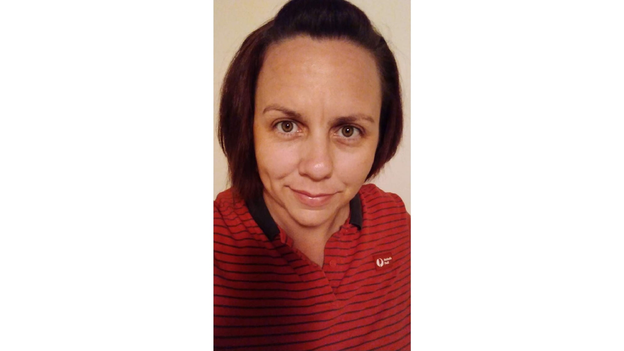 A person is shown from the shoulders up, wearing a red collared shirt with thin black horizontal stripes. The shirt features a small, clearly visible white emblem or text on the left side. The background is plain and light-colored, suggesting an indoor setting. The overall palette is dominated by red and black tones.