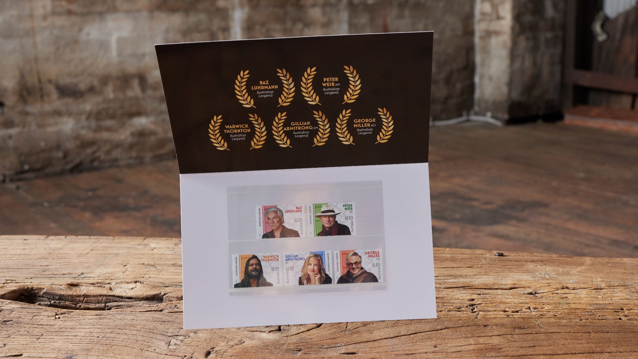A greeting card stands open on a rustic wooden table, featuring a black upper panel decorated with gold laurel wreaths and award symbols. The lower panel displays four postage stamps, each with a portrait of a different person. The background is an industrial-style room with exposed brick and wooden flooring, adding a vintage atmosphere.