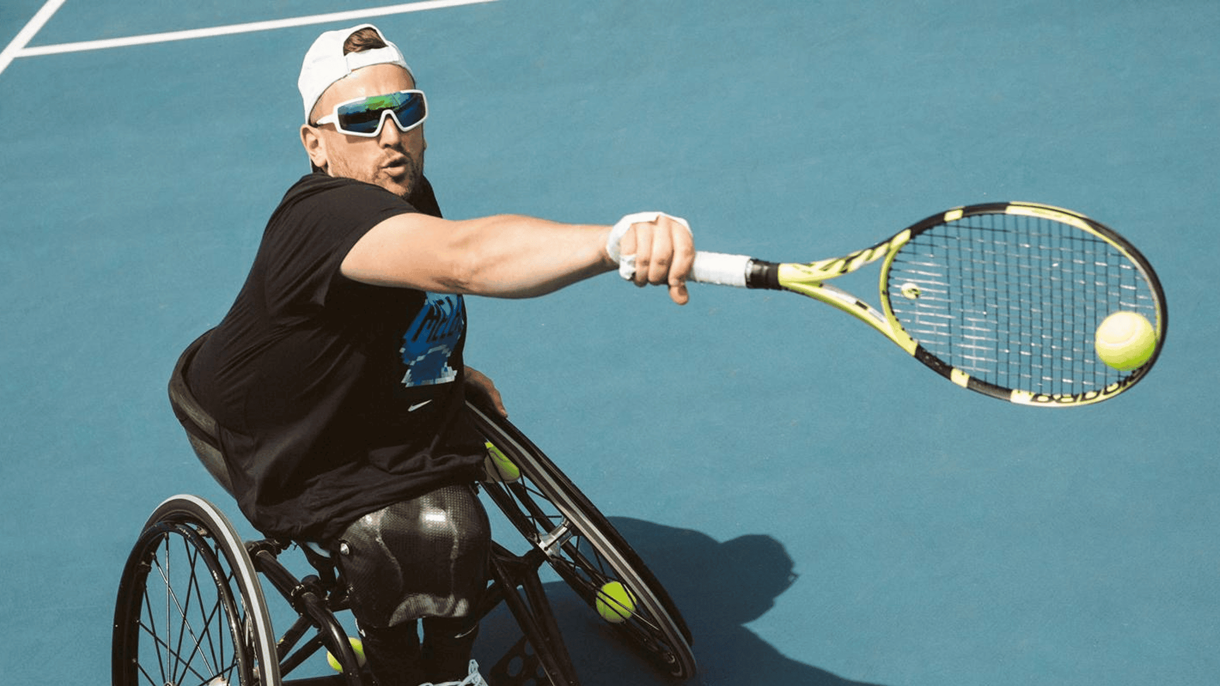 An athlete in a wheelchair is captured mid-action on a blue tennis court, swinging a tennis racquet to hit a ball. The player wears a black t-shirt and white cap, demonstrating focus and athleticism. The scene is brightly lit, emphasizing the dynamic movement and competitive spirit. The image highlights adaptive sports and inclusivity in athletics.