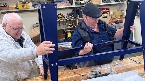 Two older men work together to assemble a blue metal frame in a workshop. They hold the structure upright on a wooden workbench surrounded by tools and hardware. Shelves with various equipment and supplies are visible in the background. The scene suggests collaborative DIY construction or furniture building in a practical, hands-on environment.