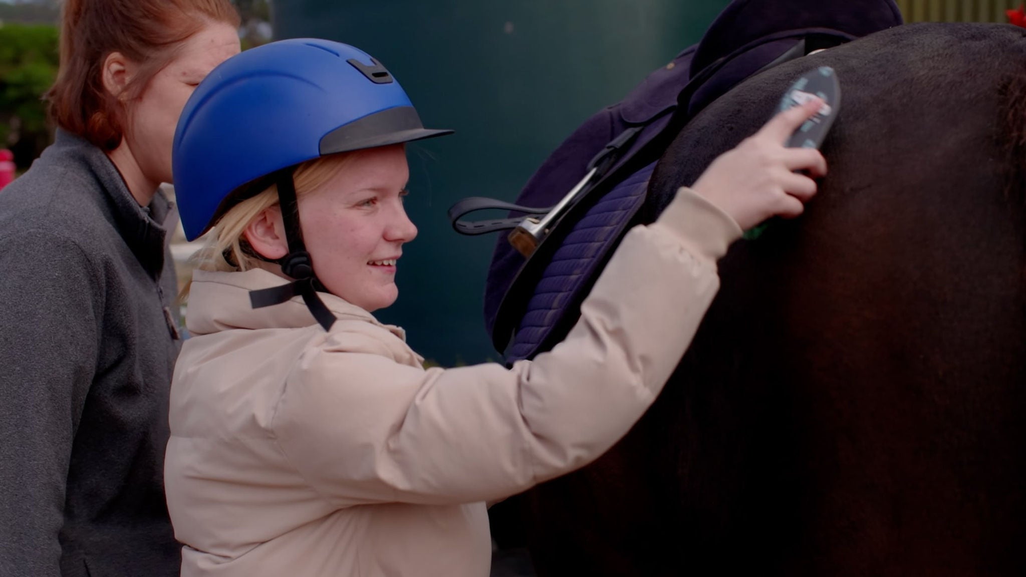 A person wearing a blue helmet and beige jacket is grooming a horse using a brush. The scene takes place outdoors, possibly at a stable or equestrian facility, with another individual present in the background. The horse is equipped with a saddle and saddle pad, and the activity appears to be part of routine horse care. The overall mood is practical and focused, with natural lighting and muted colors.