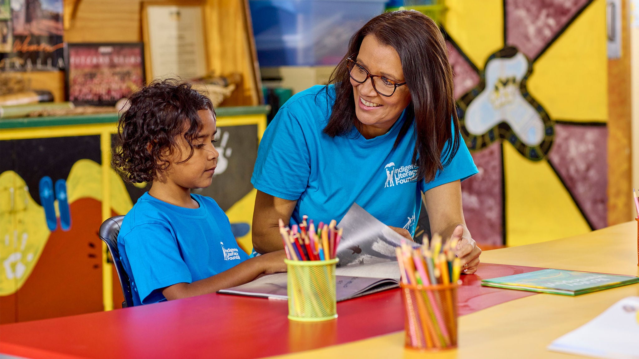 An adult and a child, both wearing matching blue t-shirts, are seated at a colorful classroom table. The adult is helping the child read a book, with colored pencils and art supplies visible on the table. The background features vibrant wall art and educational materials, creating a creative and supportive learning environment.