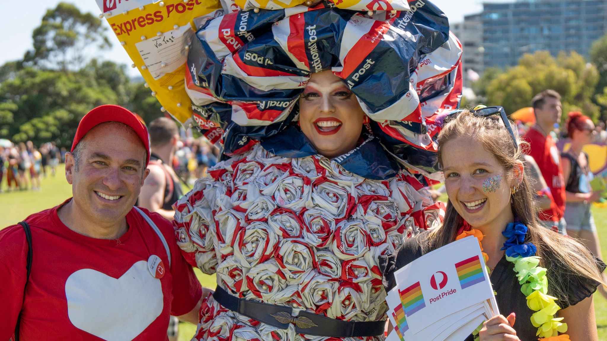 A group of people are gathered outdoors, one wearing an elaborate costume made from colorful mail packaging materials. The setting appears to be a festival or public gathering in a park, with others visible in the background. One person holds a rainbow-themed sign with 'Express Post' and 'Australia Post' clearly visible. The mood is festive and inclusive, with bright colors and creative attire.