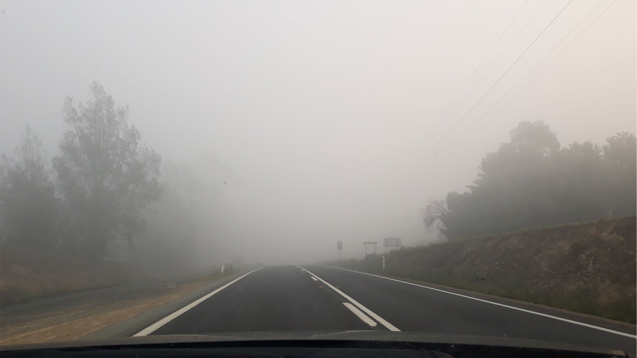 A two-lane road stretches forward, shrouded in dense fog that significantly reduces visibility. The scene is set in a rural area with trees and grassy embankments on either side. The atmosphere is muted and subdued, with soft gray tones dominating the palette. No people or vehicles are visible, emphasizing the quiet and isolated mood of the early morning drive.