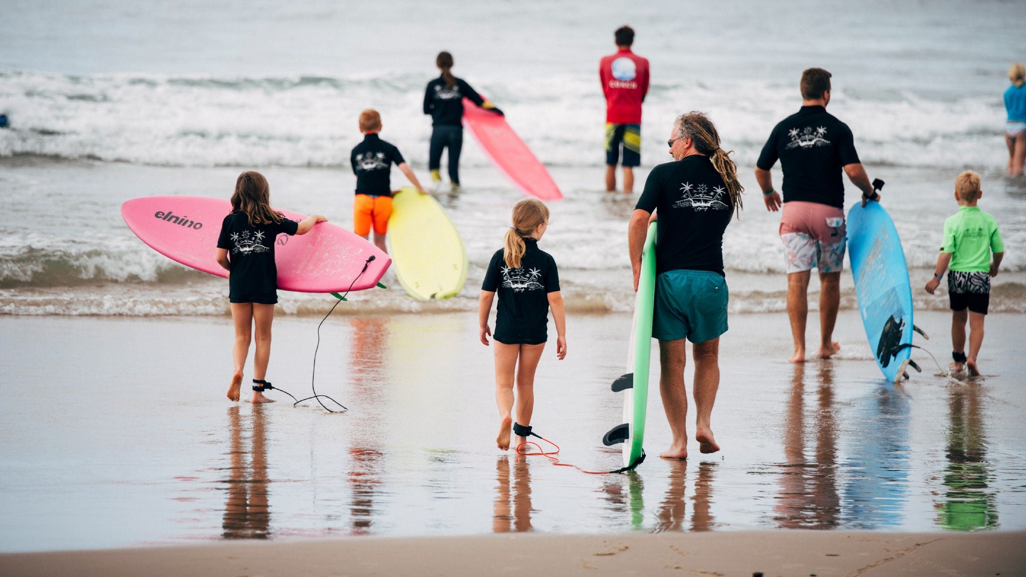 A group of children and adults, all wearing black shirts, walk toward the ocean carrying colorful surfboards. The scene takes place on a sandy beach with gentle waves in the background. The surfboards are pink, yellow, blue, and green, and one board has the word 'ohino' clearly visible. The mood is active and communal, with a focus on outdoor recreation.