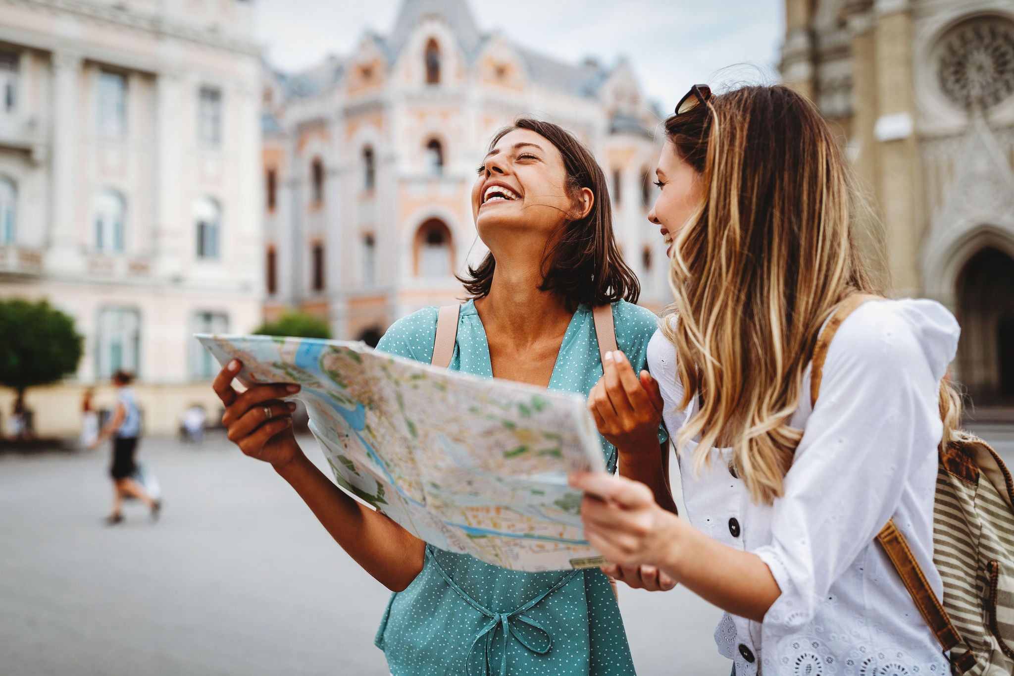 Two women are standing together in a historic city square, closely examining a paper map. The background features ornate, old European-style buildings, suggesting a travel or sightseeing activity. Both women appear to be in their twenties, dressed casually for a day of exploration. The scene conveys a sense of adventure and curiosity in an urban setting.