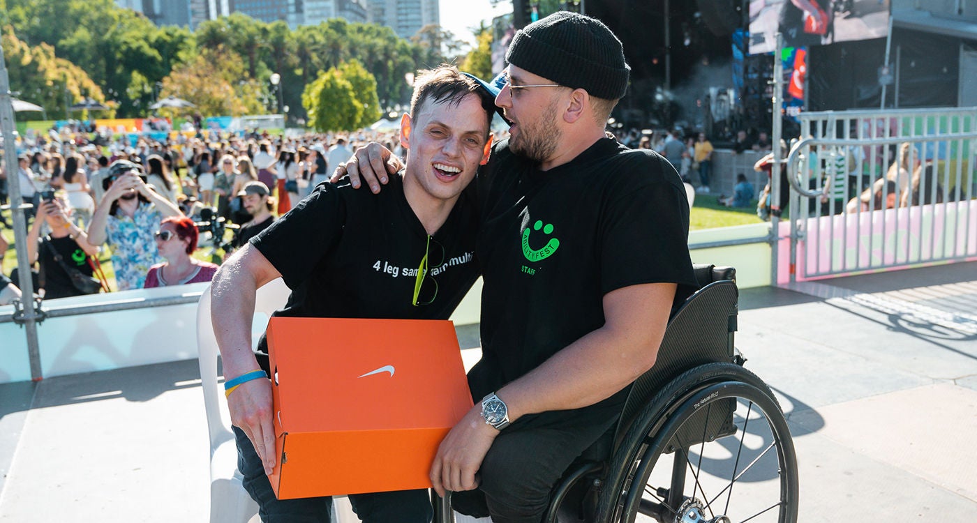 Two individuals are pictured at an outdoor event, one seated in a wheelchair and the other holding an orange Nike shoe box. The setting is a sunny public space with a crowd and stage in the background, suggesting a festival or community gathering. Both people are wearing black t-shirts with visible white and green text, and the mood is friendly and celebratory. The image captures inclusivity and positive interaction.