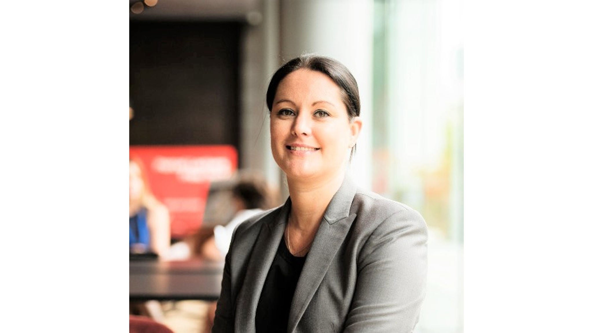 A person wearing a grey blazer and black top is seated in a contemporary office or café environment. The background features large windows with natural light and blurred figures, suggesting a collaborative or social workspace. The overall mood is professional and neutral, with a soft color palette dominated by grey and black tones. No visible text or numeric values are present in the image.
