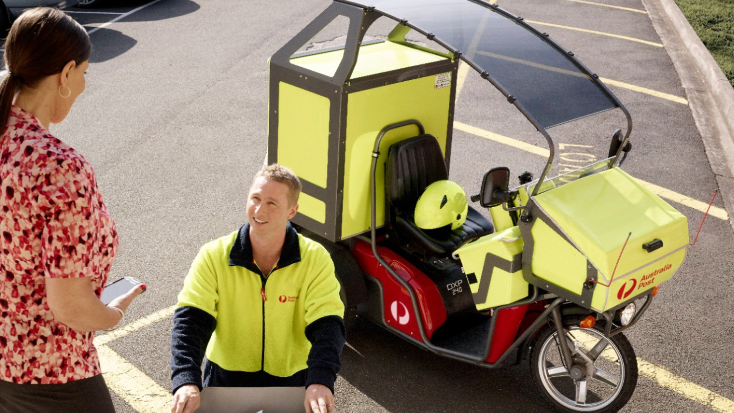 A postal delivery worker in a high-visibility jacket interacts with a woman in a parking lot. The worker is seated on a distinctive electric tricycle vehicle, which features bright yellow panels and a protective canopy. A matching yellow helmet is placed on the vehicle, and the Australia Post logo is clearly visible. The setting is a sunlit outdoor parking area with marked lines.