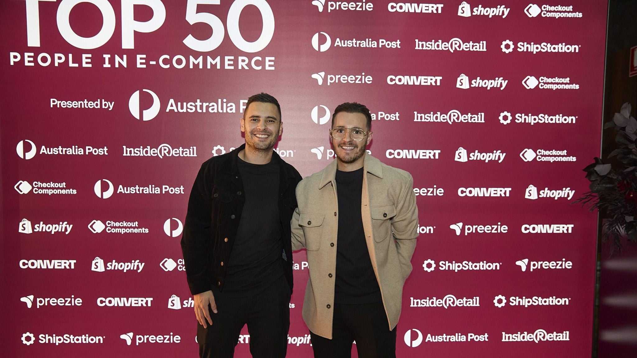 Two men stand together in front of a red step-and-repeat backdrop at an e-commerce event. The backdrop prominently features the text "TOP 50 PEOPLE IN E-COMMERCE" along with multiple sponsor names. Both individuals are dressed in smart casual attire, with jackets and dark pants, posing for a photo. The scene suggests a professional networking or awards environment focused on online retail and digital business.
