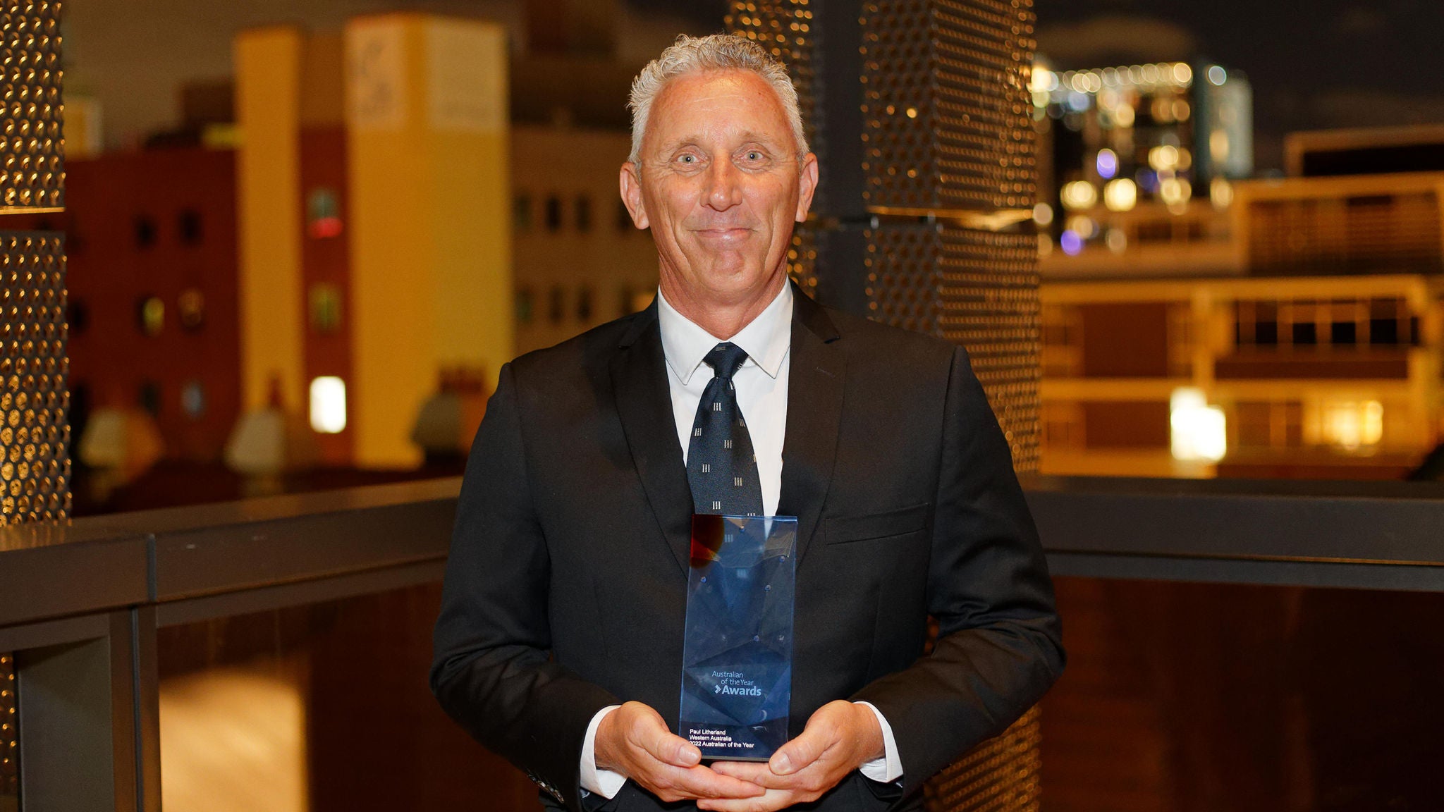 A man dressed in a formal black suit and tie stands on an outdoor balcony at night, holding a glass or crystal award. The background features illuminated city buildings and modern architecture, suggesting an urban setting. The scene conveys a celebratory or professional mood, with warm lighting and a sophisticated atmosphere. No visible text or numbers are present on the award or in the background.