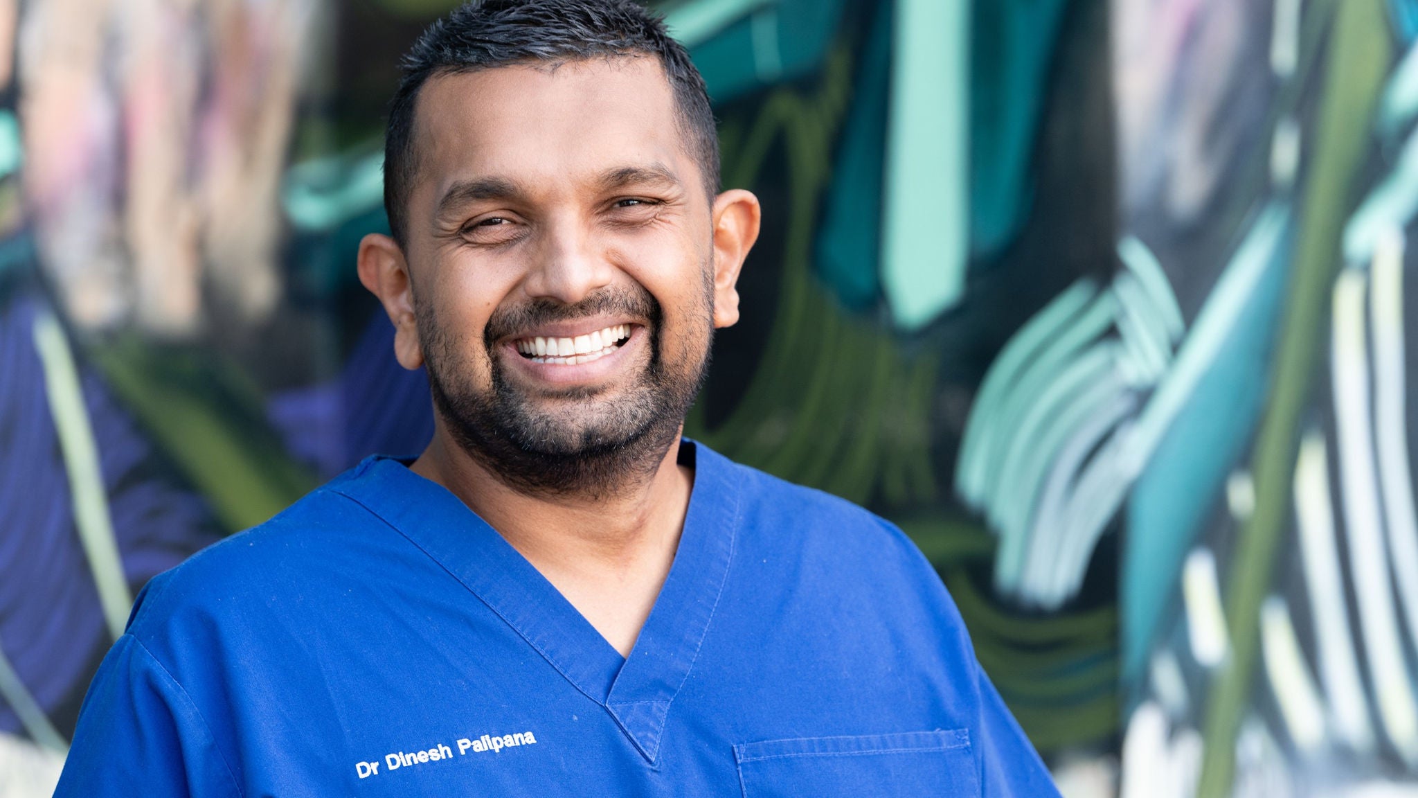 A person wearing blue medical scrubs stands in front of a colorful, abstract mural. The scrubs feature embroidered text reading 'Dr Dinesh Palipana.' The background includes bold, artistic brushstrokes in green, blue, and white tones, creating a vibrant and modern atmosphere. The subject's attire and setting suggest a healthcare or medical context.