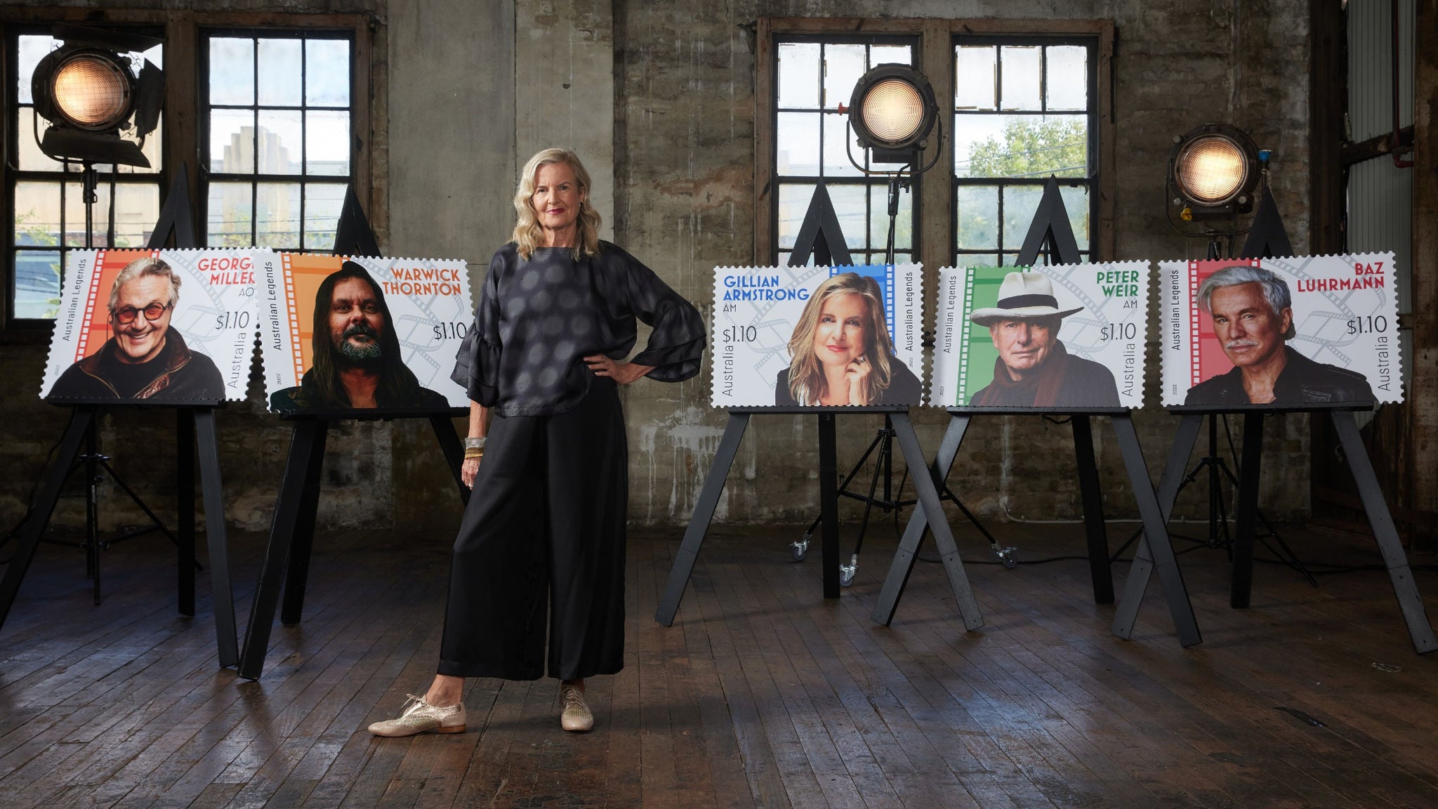 A woman stands confidently in a rustic studio space, surrounded by oversized posters designed to look like postage stamps. Each poster is mounted on an easel and features a blurred portrait with visible text and a $10 denomination. The setting includes wooden floors, industrial windows, and professional studio lighting, creating an artistic and creative atmosphere.