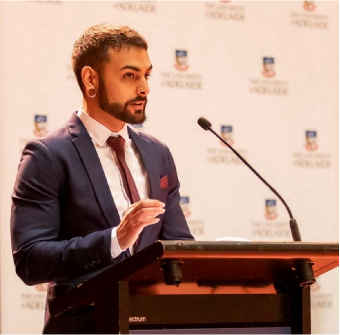 A person in formal attire stands at a podium, delivering a speech or presentation. The backdrop features repeated University of Adelaide crests and text, indicating an academic or official event. The setting is indoors with warm lighting, and the speaker is using a microphone. The overall mood is professional and focused.