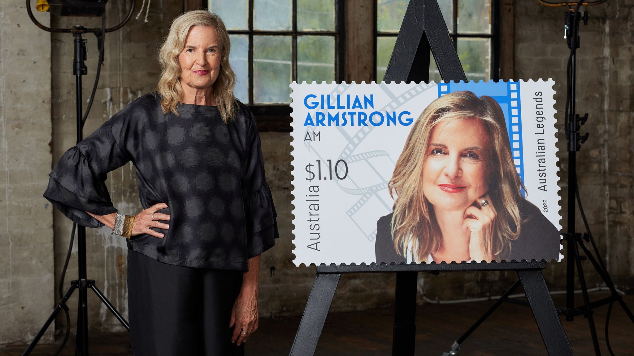 A woman stands indoors next to an oversized stamp poster displayed on an easel. The stamp features the name 'Gillian Armstrong AM,' the value '$1.10,' and the words 'Australian Legends.' The setting includes rustic windows and professional lighting equipment, creating a studio atmosphere. The woman is dressed in a dark, patterned blouse and black pants.