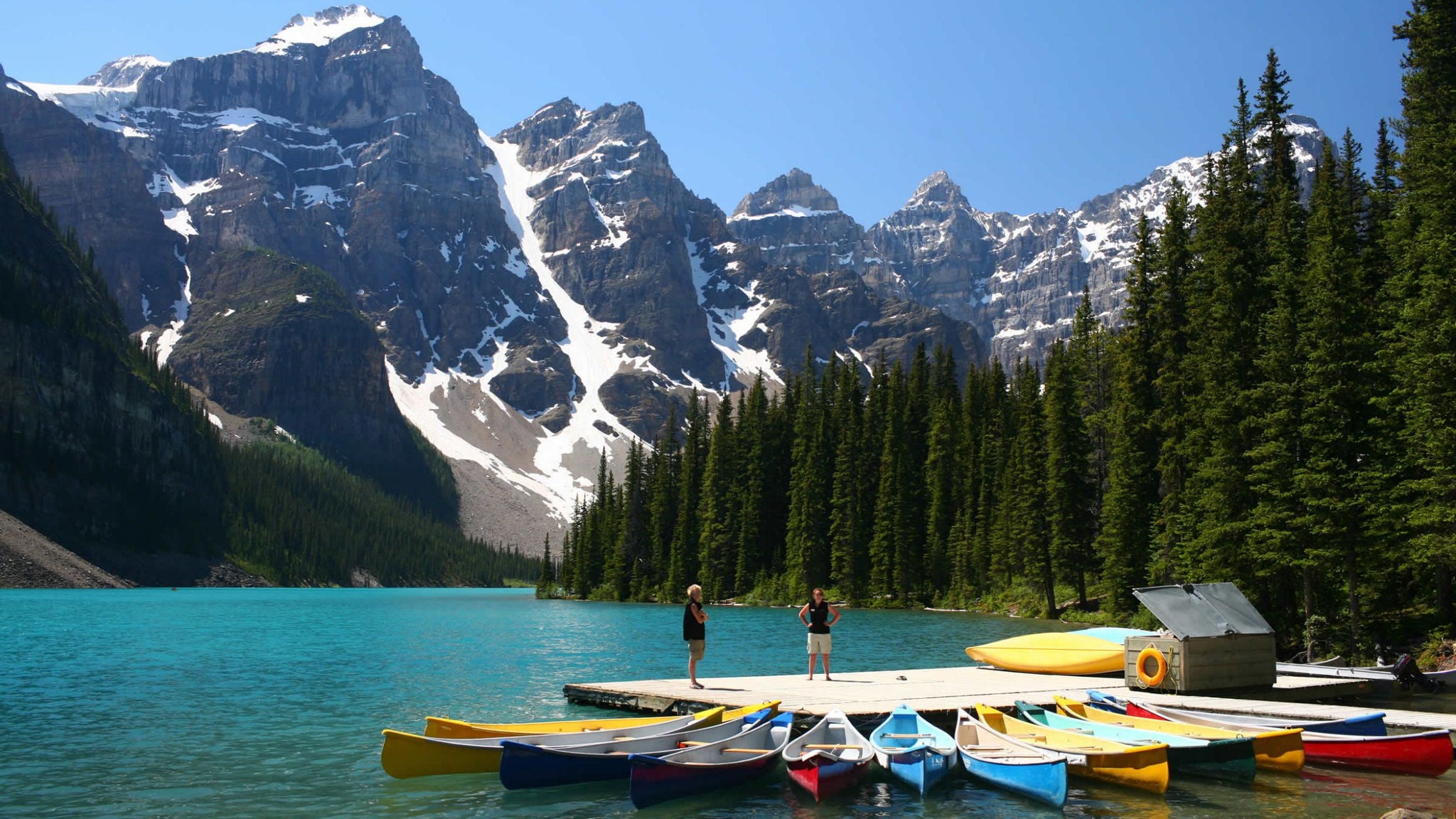 A group of colorful canoes is lined up on a wooden dock by a turquoise lake, surrounded by tall evergreen trees and dramatic snow-capped mountains. Two people stand on the dock, enjoying the scenic outdoor setting under a clear blue sky. The image captures a peaceful, adventurous mood in a natural wilderness environment.
