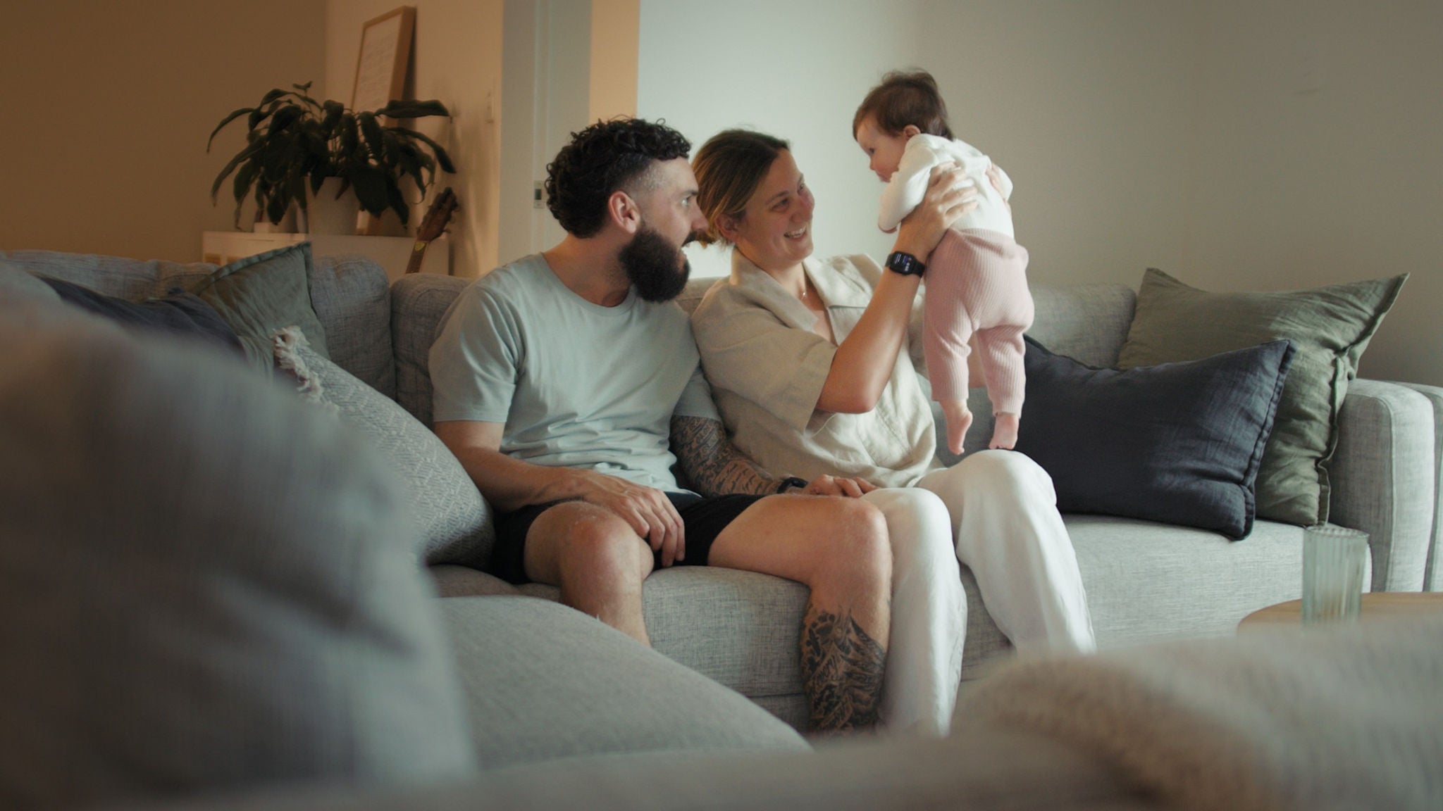 A young couple sits closely on a modern grey sofa in a cozy living room, holding a baby dressed in light pink and white. The setting features soft lighting, neutral tones, and decorative pillows, creating a warm and inviting atmosphere. A houseplant and glass of water are visible in the background, enhancing the homely feel. The scene conveys comfort, connection, and family bonding.