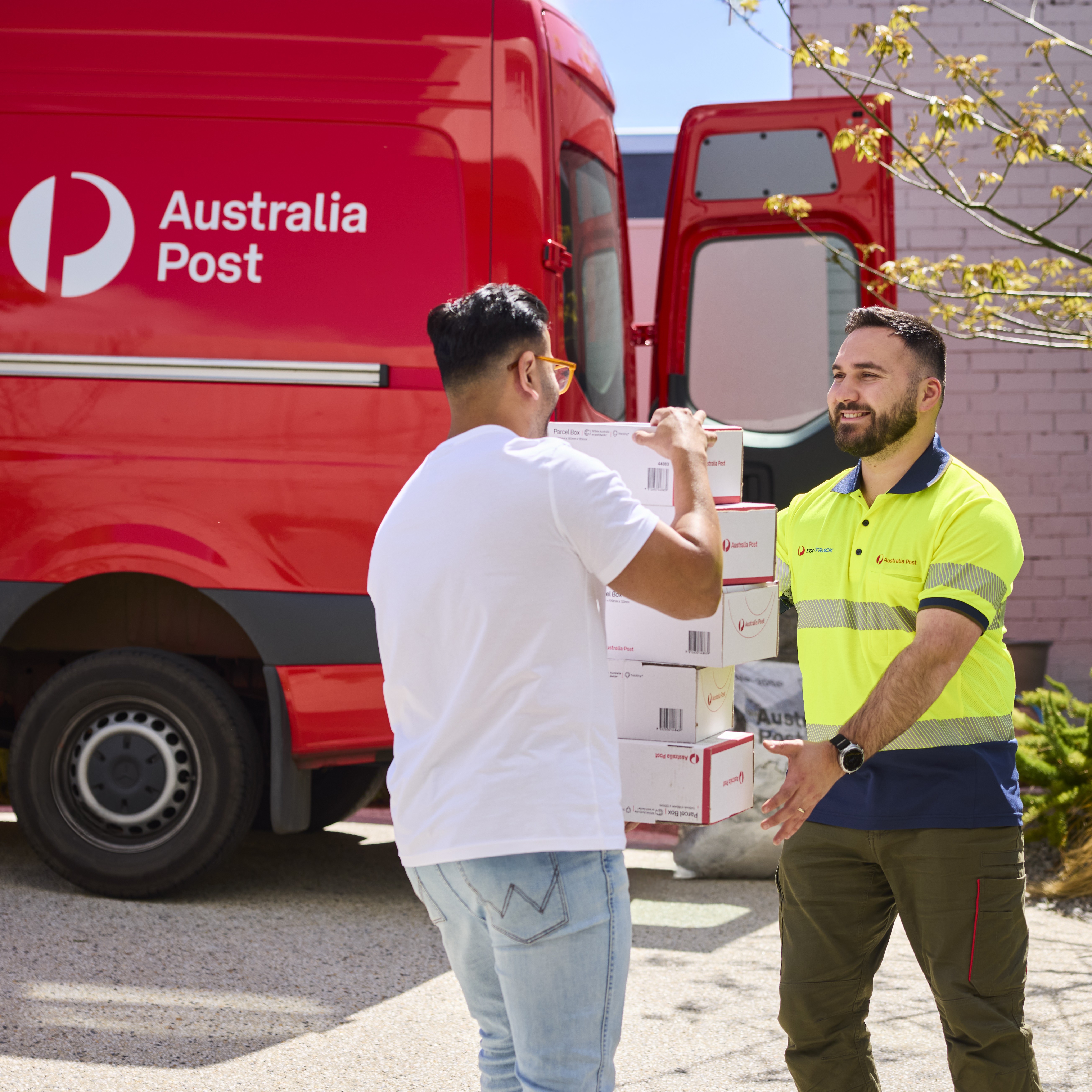 Two people transferring parcel boxes beside a red Australia Post van with its rear door open.